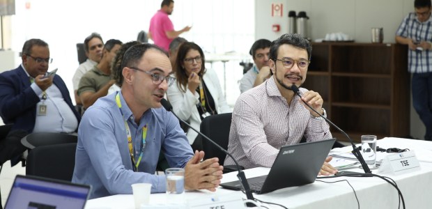 Fotografia de uma reunião em andamento, com dois participantes em destaque sentados à mesa. Um d...