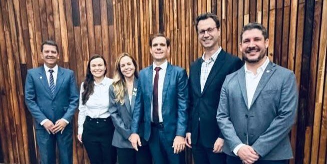Imagem de 4 homens brancos e 2 mulheres em pé, ao fundo painel em madeira.