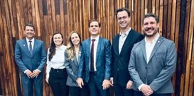 Imagem de 4 homens brancos e 2 mulheres em pé, ao fundo painel em madeira.