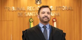 Imagem de busto de homem branco de terno e gravata com painel em madeira ao fundo e bandeiras do...