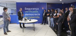 Imagem de pessoas em pé ao redor de uma mesa redonda com um homem explicando sobre o Teste públi...